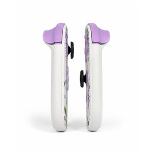 Puede incluir: Dos mandos de juego blancos colocados uno al lado del otro, con un estampado floral de flores moradas y hojas verdes. Cada mando tiene un bot&oacute;n superior de color morado claro, un joystick negro y botones direccionales morados.