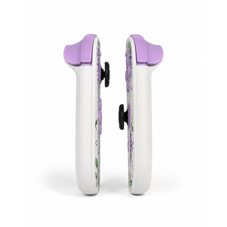 Puede incluir: Dos mandos de juego blancos colocados uno al lado del otro, con un estampado floral de flores moradas y hojas verdes. Cada mando tiene un bot&oacute;n superior de color morado claro, un joystick negro y botones direccionales morados.