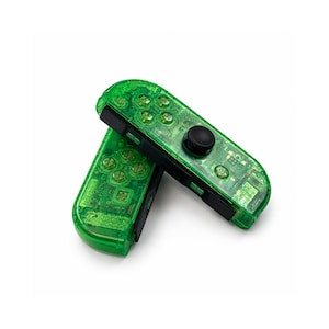 May include: Two transparent green game controllers with black accents. The controllers have visible buttons and a black joystick. The controllers are angled and overlapping, set against a white background.
