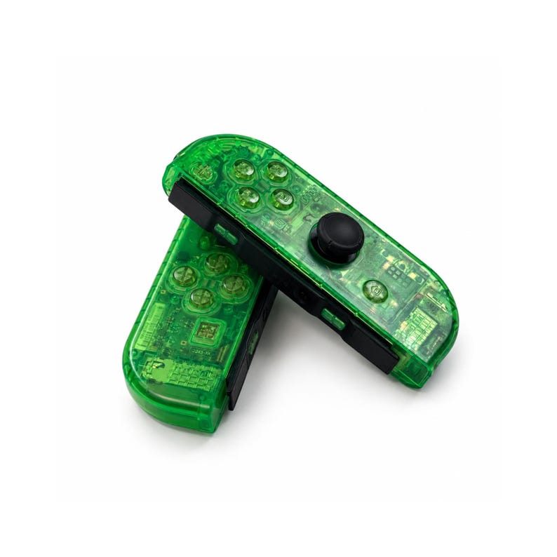 May include: Two transparent green game controllers with black accents. The controllers have visible buttons and a black joystick. The controllers are angled and overlapping, set against a white background.