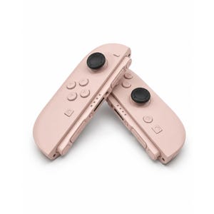 May include: Two pastel pink gaming controllers positioned at an angle. Each controller features a black circular joystick, four directional buttons, a home button, a capture button, and side-mounted shoulder buttons with a series of small indicator lights.