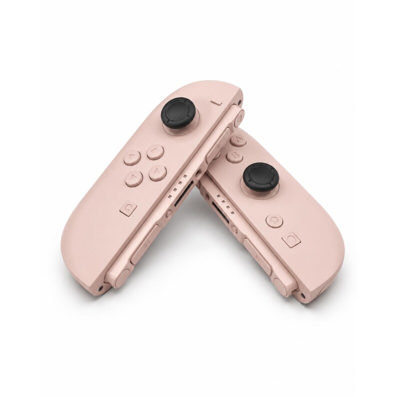 May include: Two pastel pink gaming controllers positioned at an angle. Each controller features a black circular joystick, four directional buttons, a home button, a capture button, and side-mounted shoulder buttons with a series of small indicator lights.