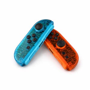 May include: A pair of wireless game controllers featuring a transparent blue and a transparent orange plastic shell. Each controller includes a black circular joystick, four directional buttons, and labeled action buttons marked A, B, X, and Y.