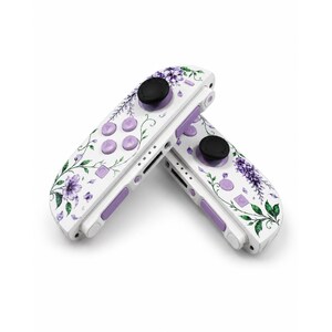 Puede incluir: Dos mandos de videojuegos blancos con un estampado floral de flores moradas y enredaderas verdes. Cada mando tiene un joystick circular negro, cuatro botones circulares de color morado claro y un bot&oacute;n lateral rectangular.