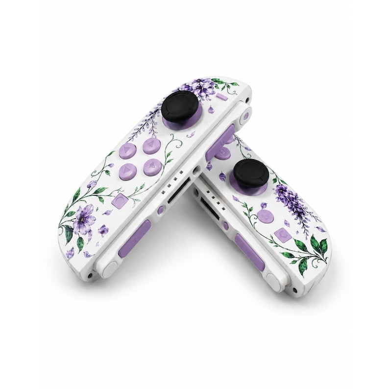 Puede incluir: Dos mandos de videojuegos blancos con un estampado floral de flores moradas y enredaderas verdes. Cada mando tiene un joystick circular negro, cuatro botones circulares de color morado claro y un bot&oacute;n lateral rectangular.