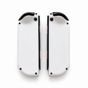 Pode incluir: Dois controles de jogo brancos e pretos, cada um com uma parte superior preta, bot&otilde;es pretos e detalhes em vermelho. Os controles est&atilde;o posicionados lado a lado em um fundo branco.