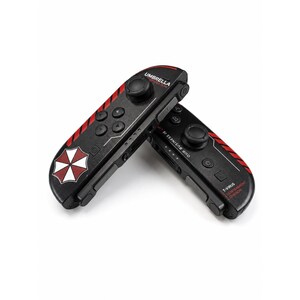 May include: Two black video game controllers featuring red diagonal stripes and the Umbrella Corporation logo. The controllers include buttons, a joystick, and text that reads Umbrella Corporation, Our Business Is Life Itself, and T-Virus.