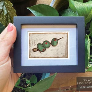 May include: A framed painting of a green olive skewer on a white background. The olives are painted in a simple, stylized manner. The frame is black with a white mat.