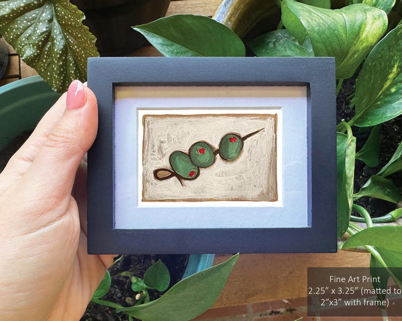 May include: A framed painting of a green olive skewer on a white background. The olives are painted in a simple, stylized manner. The frame is black with a white mat.