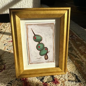 May include: A framed painting of a green olive branch with four olives. The olives are painted in a realistic style with a green hue and red centers. The painting is framed in a gold frame.