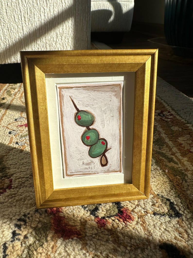 May include: A framed painting of a green olive branch with four olives. The olives are painted in a realistic style with a green hue and red centers. The painting is framed in a gold frame.