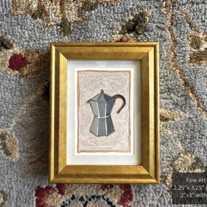 May include: A framed print of a silver coffee pot with a black handle on a light pink background. The print is 2.25 inches by 3.25 inches and is matted to 2 inches by 3 inches with a gold frame.