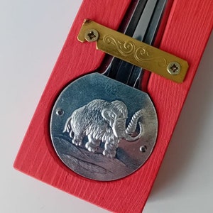 Op de afbeelding: Een rode houten doos met een metalen mammoetontwerp aan de voorkant. De doos bevat een metalen instrument met een messinghaak.