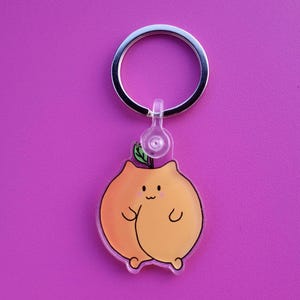 May include: A peach-shaped keyring with a cute cartoon cat face and a green leaf on top. The keyring is made of clear acrylic and has a silver metal ring.