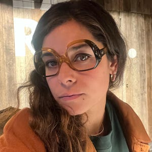 May include: Large, round eyeglasses with a brown and black frame. The lenses are clear. The person is wearing an orange jacket and a green shirt. The word "EAT" is visible in white letters on a wooden background.