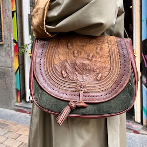 May include: A brown leather satchel bag with a green suede bottom. The bag has a flap with a tassel and is decorated with a floral design.