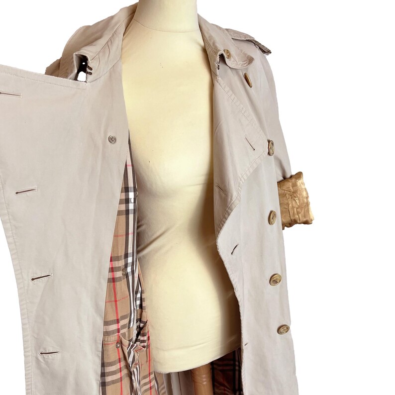 Puede incluir: Gabardina beige con un dise&ntilde;o cl&aacute;sico. El abrigo presenta un frente cruzado, cierres de botones y un cuello. El forro interior muestra un estampado de cuadros. El abrigo se exhibe en un maniqu&iacute;.