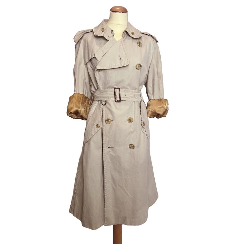 Puede incluir: Un trench coat beige con dise&ntilde;o cruzado, cintur&oacute;n en la cintura y pu&ntilde;os dorados. El abrigo se exhibe en un maniqu&iacute; y presenta un estilo cl&aacute;sico y atemporal. La longitud del abrigo es de aproximadamente 114 cm.