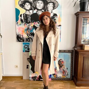 Puede incluir: Un abrigo beige con dise&ntilde;o de doble botonadura y boina marr&oacute;n. El abrigo se lleva sobre un vestido negro y se combina con botines negros. El fondo presenta obras de arte coloridas y un suelo de madera.
