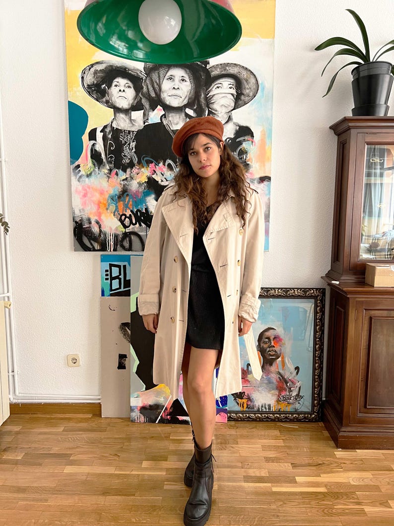 Puede incluir: Un abrigo beige con dise&ntilde;o de doble botonadura y boina marr&oacute;n. El abrigo se lleva sobre un vestido negro y se combina con botines negros. El fondo presenta obras de arte coloridas y un suelo de madera.