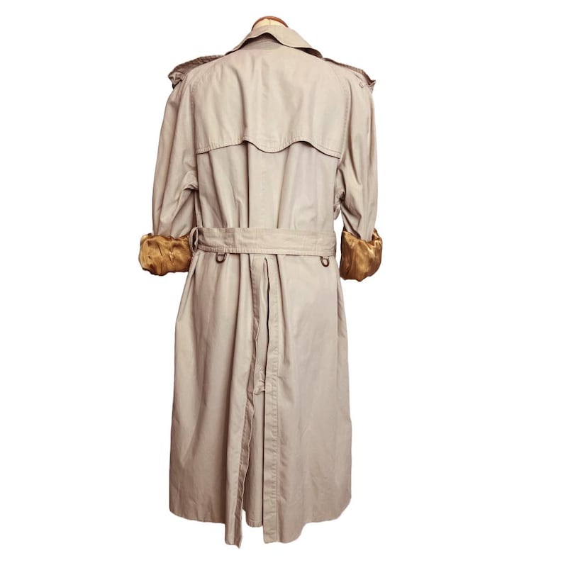 Puede incluir: Gabardina beige con cintur&oacute;n en la cintura y charreteras decorativas. El abrigo presenta un dise&ntilde;o cl&aacute;sico con doble botonadura y pu&ntilde;os de piel sint&eacute;tica dorada. Una prenda de abrigo atemporal.