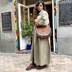 May include: A woman wearing a long beige trench coat with a brown leather satchel bag. She is standing on a brick sidewalk in front of a building with a chalkboard menu that says "HOMEMADE FOOD FOR THE SOUL".
