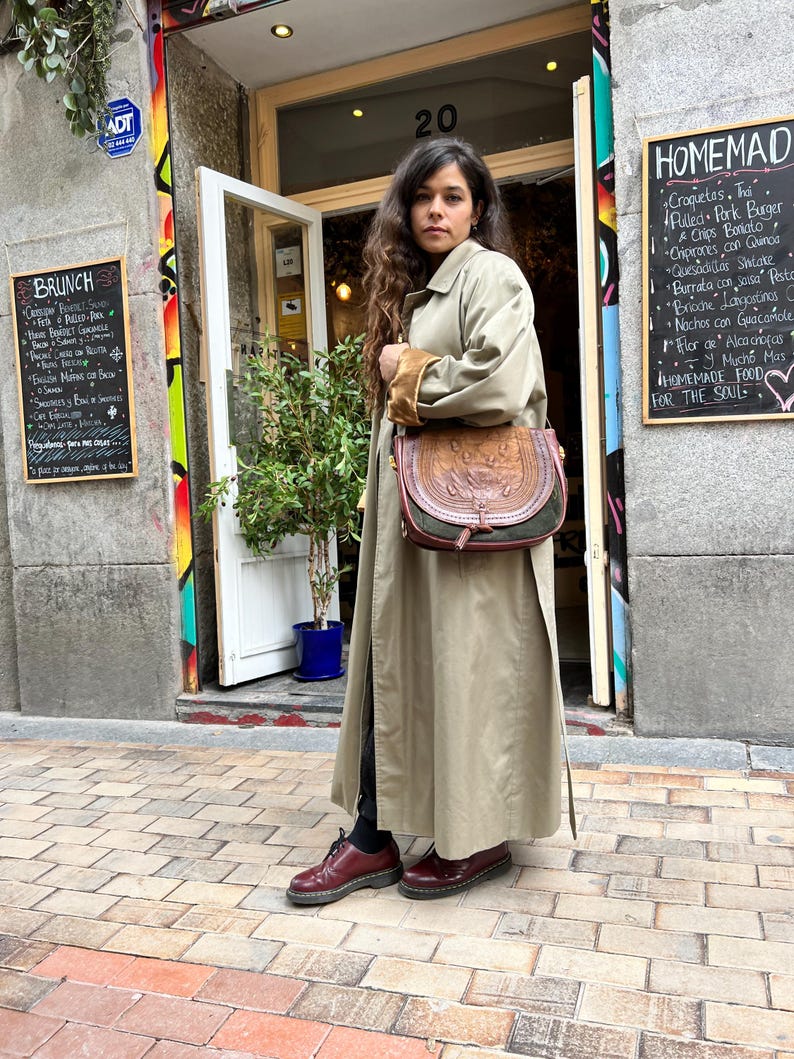 May include: A woman wearing a long beige trench coat with a brown leather satchel bag. She is standing on a brick sidewalk in front of a building with a chalkboard menu that says "HOMEMADE FOOD FOR THE SOUL".