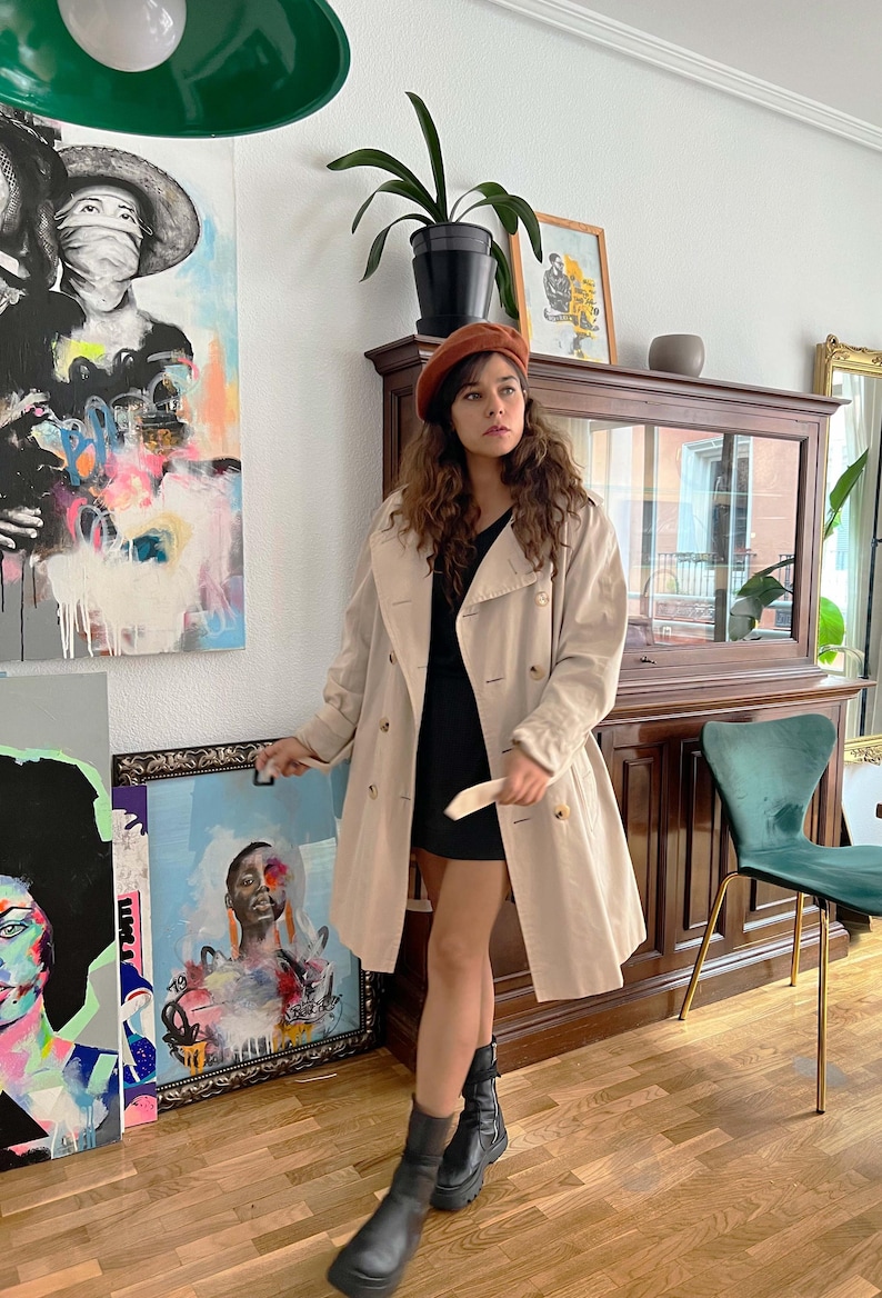 Puede incluir: Un abrigo tipo trench beige con dise&ntilde;o de doble botonadura. Se lleva sobre un vestido negro y se combina con botas negras. Se lleva una boina marr&oacute;n en la cabeza. El fondo incluye obras de arte y un mueble de madera.