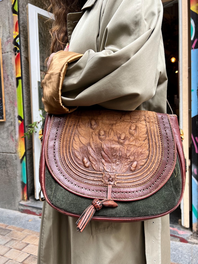 May include: A brown leather satchel bag with a green suede bottom. The bag has a flap with a tassel and is decorated with a floral design.