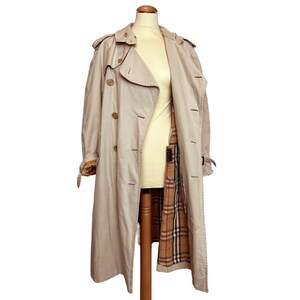 Puede incluir: Un trench coat beige con un dise&ntilde;o cl&aacute;sico. El abrigo presenta una parte delantera cruzada, cierres de botones y una cintura con cintur&oacute;n. El forro interior muestra un estampado de cuadros en tonos marrones, rojos y negros. El abrigo se exhibe en un maniqu&iacute;.