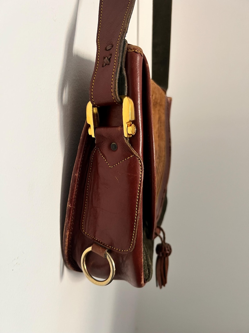 May include: A brown leather shoulder bag with a gold buckle and a tassel. The bag has a flap with a small pocket and a ring on the side.