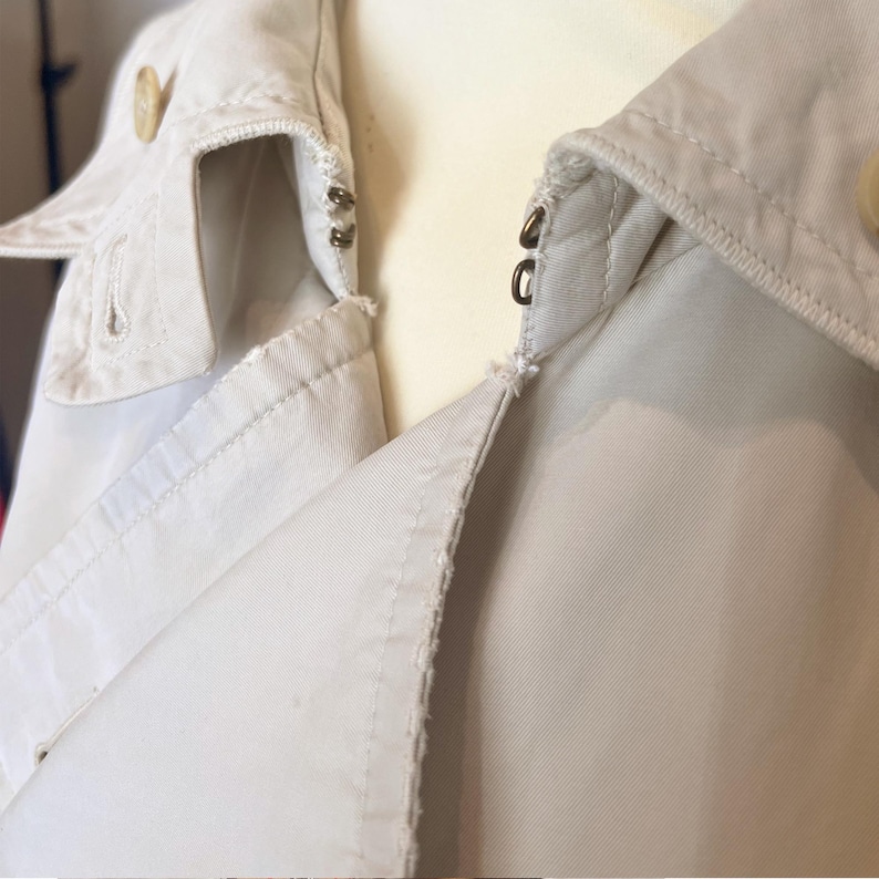 Puede incluir: Primer plano de un cuello de chaqueta beige con cierre de bot&oacute;n. La chaqueta tiene un dise&ntilde;o cl&aacute;sico con solapas y cierre de botones en la parte delantera. La tela parece ser un material ligero, adecuado para la primavera o el oto&ntilde;o.