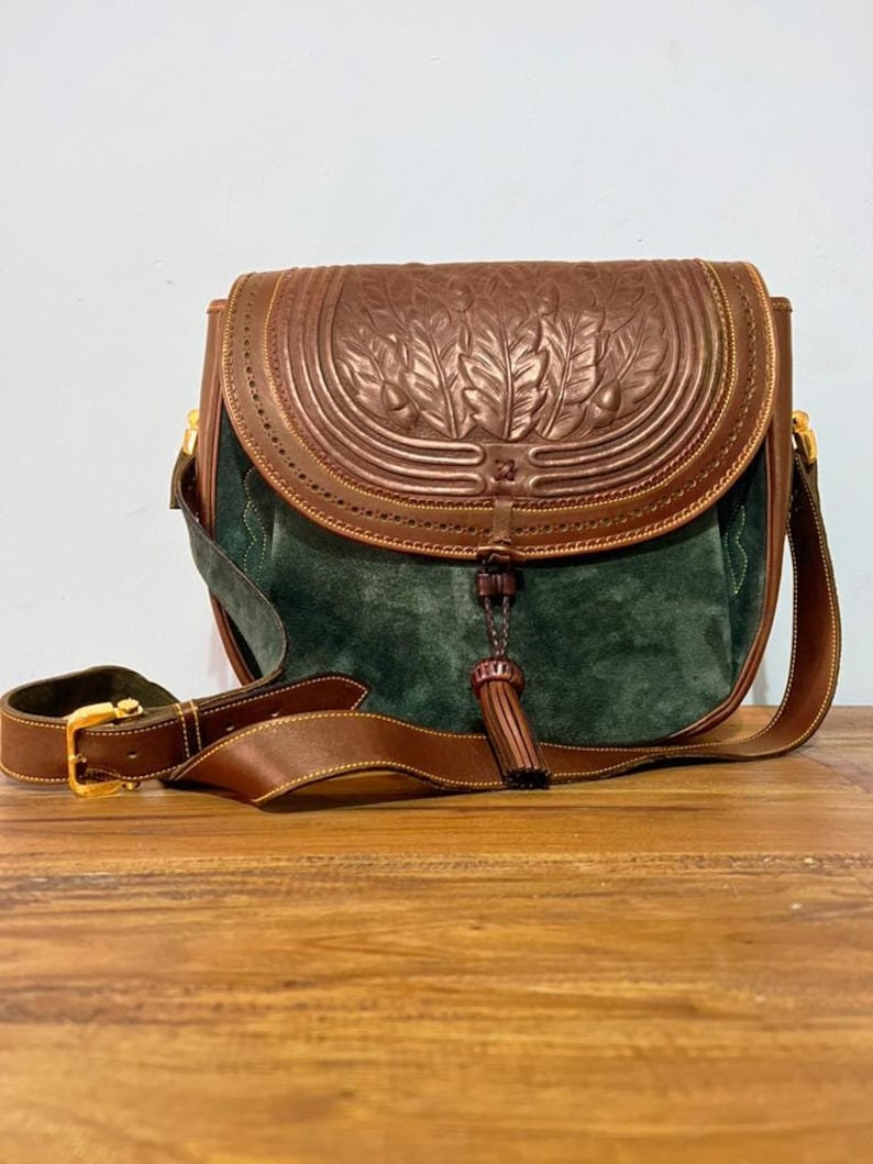 May include: A brown leather shoulder bag with a green suede flap and a tassel. The bag has a decorative embossed design of leaves and acorns.