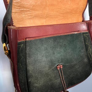 May include: A dark green suede shoulder bag with a burgundy leather flap and trim. The bag has a gold-tone buckle closure and a tassel.
