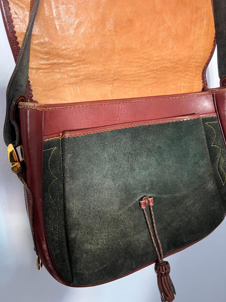 May include: A dark green suede shoulder bag with a burgundy leather flap and trim. The bag has a gold-tone buckle closure and a tassel.