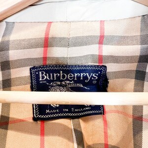 Puede incluir: Primer plano del forro beige de una chaqueta Burberry con un patr&oacute;n de cuadros cl&aacute;sico en beige, rojo y negro. Se muestra una etiqueta azul marino con el logotipo de Burberry y el texto "Made in England".