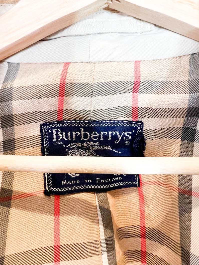 Puede incluir: Primer plano del forro beige de una chaqueta Burberry con un patr&oacute;n de cuadros cl&aacute;sico en beige, rojo y negro. Se muestra una etiqueta azul marino con el logotipo de Burberry y el texto "Made in England".