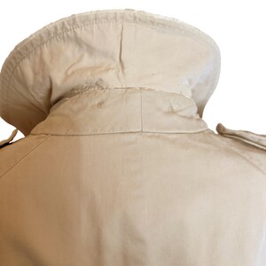 Puede incluir: Primer plano del cuello y el hombro de una gabardina beige. El cuello es de forma redondeada y la gabardina parece estar hecha de una tela resistente. El hombro tiene una charretera decorativa. El fondo es blanco.