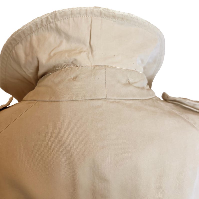 Puede incluir: Primer plano del cuello y el hombro de una gabardina beige. El cuello es de forma redondeada y la gabardina parece estar hecha de una tela resistente. El hombro tiene una charretera decorativa. El fondo es blanco.