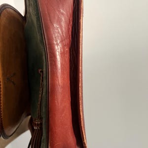 May include: A brown leather and green suede shoulder bag with a tassel. The bag has a flap closure and a gold metal ring.