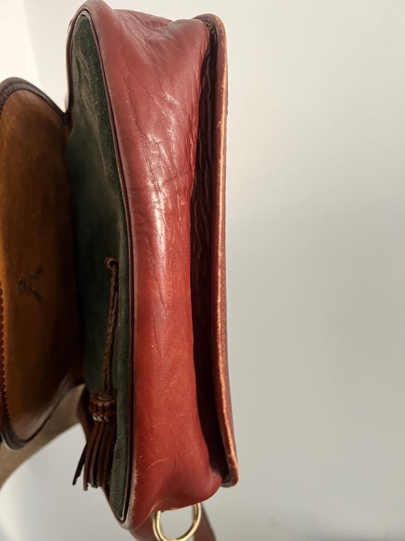 May include: A brown leather and green suede shoulder bag with a tassel. The bag has a flap closure and a gold metal ring.