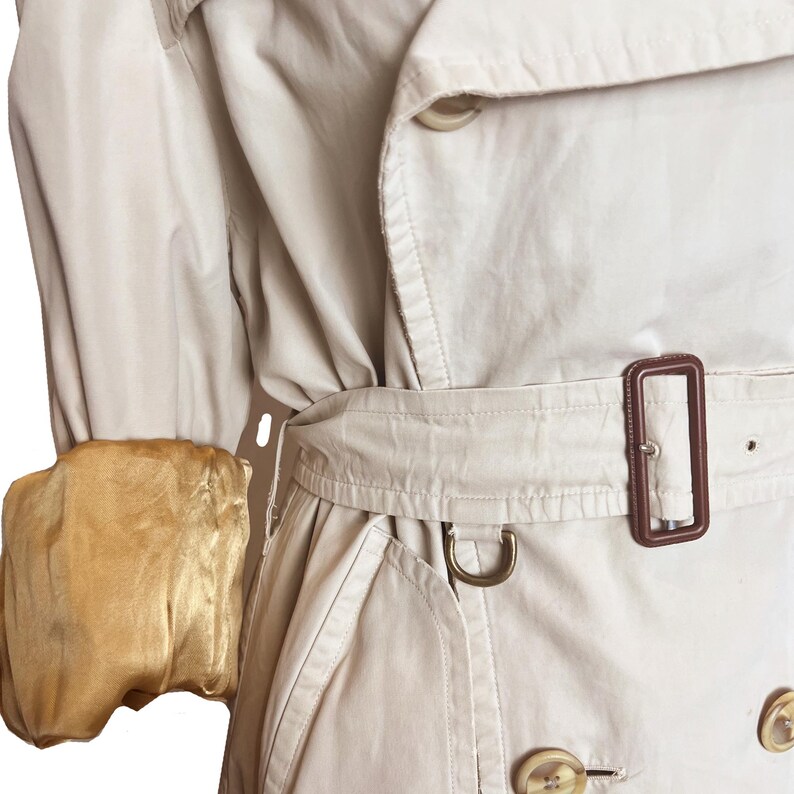 Puede incluir: Primer plano de una gabardina beige. El abrigo presenta un dise&ntilde;o cruzado con botones grandes, un cintur&oacute;n con hebilla marr&oacute;n y un pu&ntilde;o con forro dorado. El abrigo est&aacute; hecho de una tela lisa.