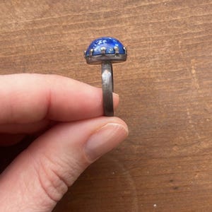 Puede incluir: Un anillo de plata con una piedra azul grande y redonda. La piedra tiene un aspecto moteado. El anillo se sostiene sobre un fondo de madera.