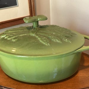May include: A green cast iron Dutch oven with a matching lid. The lid features a wheat stalk design and a stem-shaped handle. The pot sits on a wooden table. A framed picture hangs in the background.