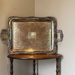 May include: A rectangular silver-plated serving tray with ornate, embossed floral detailing along the raised edges and two decorative handles. The center of the tray features an intricate, engraved pattern and a smooth, rectangular central plaque.