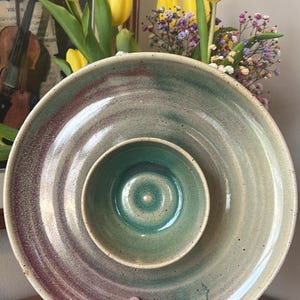 以下が含まれることがあります： 手作りのセラミック製サービング皿。同心円状のデザインで、外側のリングはくすんだ緑色にマルーンの帯、中央のボウルは鮮やかなターコイズ色です。黄色いチューリップを背景に、この皿が手に持たれています。