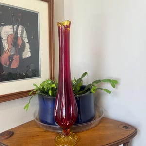 May include: A tall, slender glass vase featuring a deep red body that transitions into a vibrant amber yellow base. The vase has a flared, organic rim and a vertical ribbed texture along its elongated neck, standing upright on a circular foot.