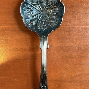 May include: Silver serving spoon with a decorative floral design on the bowl and handle. The bowl has a scalloped edge and is adorned with detailed fruit and leaf patterns. The handle features an ornate, engraved design.