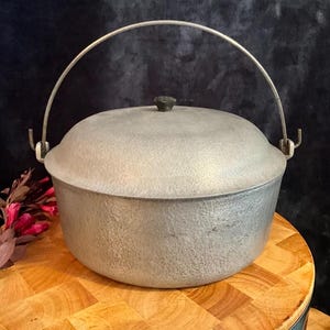 Könnte beinhalten: Ein runder, silberfarbener Dutch Oven aus Metall mit strukturierter Außenfläche. Der Topf verfügt über einen passenden gewölbten Deckel mit einem kleinen, dunklen Mittelknopf und einen geschwungenen Metallbügel, der an zwei Seiten befestigt ist.
