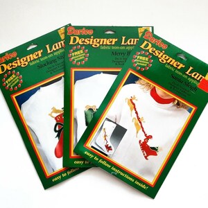 Op de afbeelding: Drie Darice Designer Lane strijkapplicatiepakketten. De pakketten zijn groen met rode en gele accenten. Elk pakket heeft een ander kerstontwerp, waaronder een kous, een slee en een cadeau.